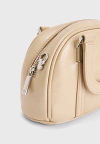 Beige leren handtas met zilveren rits en sluiting, gebogen ontwerp met gestikte details en een omhulde handgreep.