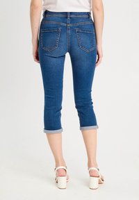 Jean capri en denim bleu avec ourlets retroussés, comprenant deux poches arrière et une coupe ajustée. Le tissu a une texture lisse et un lavage moyen.