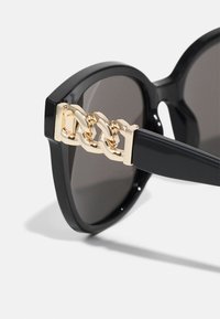 Black sunglasses with a chunky frame, featuring gray lenses and a gold chain accent on the temple. Smooth glossy finish.