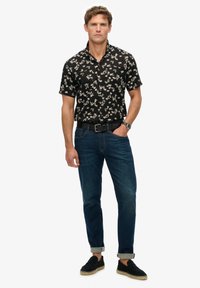 Camisa negra de manga corta con patrón floral blanco, combinada con jeans azul oscuro y zapatos de slip-on negros, mostrando un ajuste relajado.