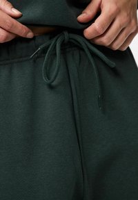 Ciemnozielony materiał bluzy z kapturem z regulowanym paskiem w talii, charakteryzujący się gładką fakturą i minimalnymi detalami szwów. Casualowy design z luźnym krojem.