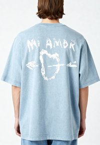 T-shirt oversized di colore blu chiaro con maniche corte. Sul retro presenta un testo grafico bianco "Mi Amor" e un design con cuore e freccia. Materiale morbido in cotone.