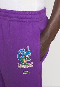 Close-up af en person iført lilla joggingbukser med elastisk taljebånd, med et "Club Lacoste"-logo og en grøn krokodilleemblem.