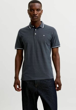 Man met een donkergrijs poloshirt met witte bies aan kraag en mouwen en een donkere broek, staand tegen een effen lichte achtergrond.