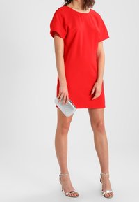 Červené šaty s krátkým rukávem a volným střihem, hladká textura. Modelka drží stříbrný psaníčko se třpytkami a má na sobě stříbrné sandály na vysokém podpatku.