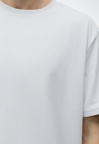 Κοντινή λήψη ενός ατόμου που φοράει μια απλή λευκή κοντομάνικη μπλούζα με ορατές ραφές στο τελείωμα του μανικιού.
