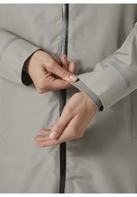 Chaqueta impermeable gris claro con cremallera, que presenta un diseño de puños en contraste. Manos ajustando la manga para un mejor ajuste, mostrando una textura suave.