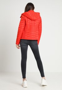 Femme portant une veste matelassée rouge vif à capuche, un jean slim foncé et des baskets blanches épaisses, debout de dos sur un fond uni.