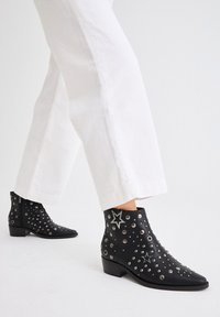 Botas de tobillo de cuero negro con tachuelas plateadas y acentos de estrellas, con un tacón bajo y un diseño de punta afilada.