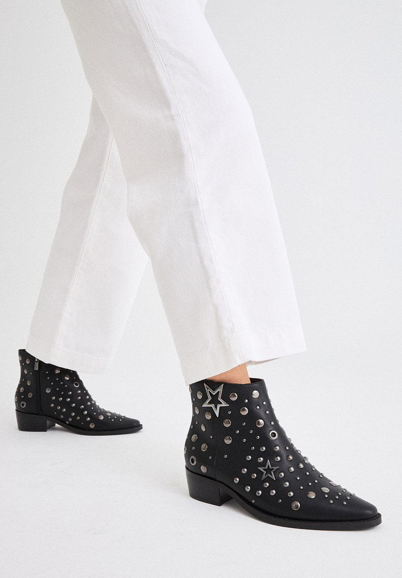 Botas de tobillo de cuero negro con tachuelas plateadas y acentos de estrellas, con un tacón bajo y un diseño de punta afilada.