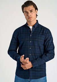 Jonge man in een donkerblauwe geblokte overhemddas met een wit T-shirt eronder, die zijn manchet aanpast terwijl hij tegen een effen achtergrond staat.