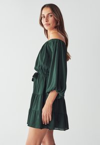 Dunkelgrünes, schulterfreies Kleid mit Ballonärmeln, tailliertem Bund und gestuftem Rock. Aus leichtem, strukturiertem Stoff gefertigt.