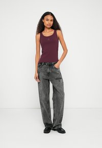 Bordeauxrotes Tanktop mit Rundhalsausschnitt und kleinem Logo. Lockere, ausgewaschene schwarze Jeans und schwarze Schuhe mit strukturierten Akzenten.