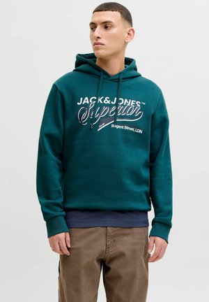 Jeune homme portant un sweat à capuche bleu sarcelle avec le texte "Jack & Jones Superior", regardant sur le côté, avec un pantalon brun et une chemise bleu marine en dessous.