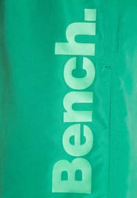 Zelená tkanina s veľkým, výrazným bielym logom "Bench." na boku. Materiál sa zdá byť hladký s našitými detailami.