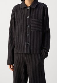 Set di abbigliamento da casa a righe nere, realizzato in tessuto morbido, composto da una giacca con colletto, cinque bottoni e taschino sul petto, abbinata a pantaloni coordinati.