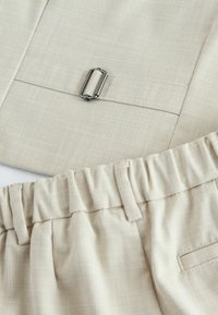 Nahaufnahme von beigefarbenen Stoffhosen mit elastischem Bund und verstellbarer Metallschnalle an passendem Stoffgürtel.