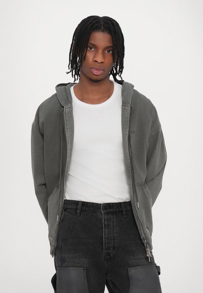 Haut à capuchon zippé gris foncé avec une capuche doublée, porté sur un t-shirt blanc. Associé à un jean noir avec des genouillères et une coupe décontractée.