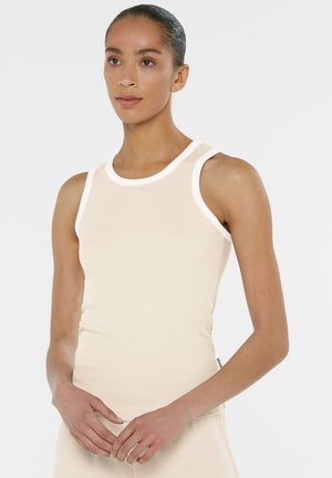 Woman with tied-back hair wearing a sleeveless beige top with white trim, standing with hands relaxed in front against a white background.