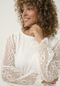 White lace top featuring diamond patterns, puffed long sleeves with ruffled cuffs, and intricate detailing throughout the fabric.