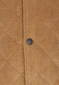 Barázdált kordbársony anyag bézs színben, függőleges bordázattal. A szövet közepén elhelyezett fém gomb található, amely a márkát azonosítja.