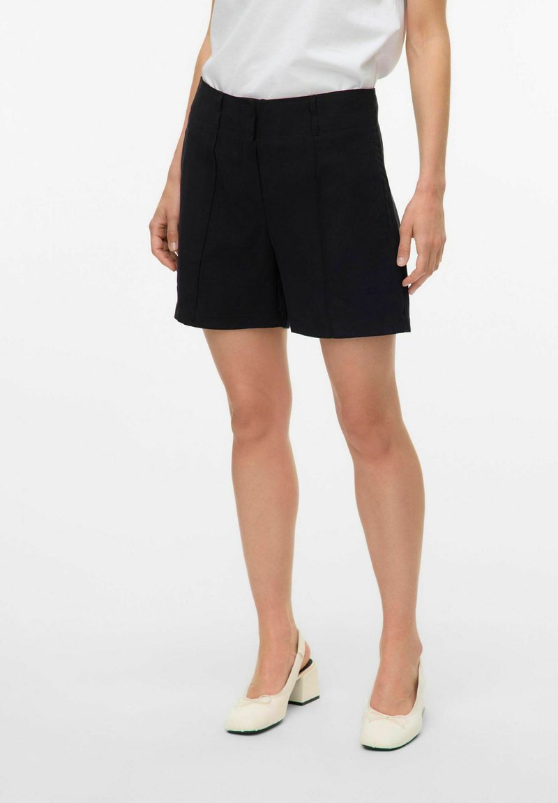 Zwarte op maat gemaakte shorts met een gladde textuur, voorzien van voorplooien en een tailleband met een gemiddelde hoogte; gecombineerd met witte, lage hak schoenen.