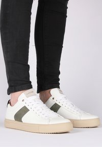 Zapatillas blancas con un acabado texturizado, que cuentan con una franja de acento verde, suela de goma beige y sistema de cordones. Etiqueta de la marca visible en la lengüeta.