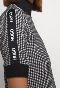 Sort-hvid houndstooth mønstret striktrøje med høj hals, der har sorte ribbede manchetter og tre hvide "HUGO" logoer på ærmerne.