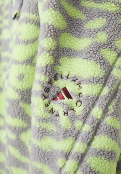 Fazenda de felpa verde e cinza com uma textura peluda, apresentando um logótipo circular bordado em vermelho e um fecho vertical.