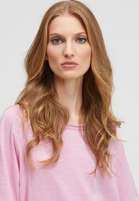 Woman with long wavy light brown hair, wearing a light pink top, facing forward with a neutral expression against a plain light background.