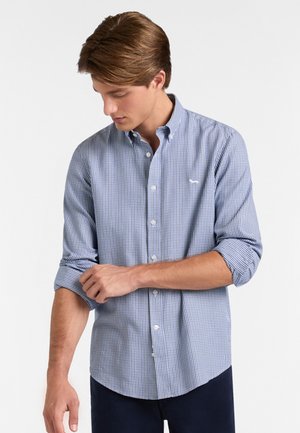 Joven de pie contra un fondo blanco liso, enrollando las mangas de una camisa de cuadros azul y blanca.