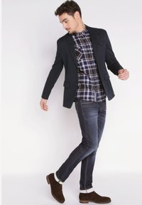 Laivastonsininen bleiseri musta-valkoisen ruudullisen paidan päällä, yhdistetty tummiin farkkuihin ja ruskeisiin mokkasiineihin, joissa on käärityt hihat ja räätälöity istuvuus.