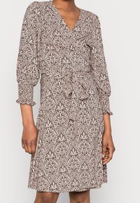 Mujer vistiendo un vestido envolvente beige hasta la rodilla con un intrincado patrón floral negro y mangas tres cuartos recogidas, atado en la cintura.