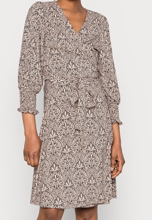 Femme portant une robe portefeuille beige midi avec un motif floral noir complexe et des manches rassemblées à 3/4, nouée à la taille.