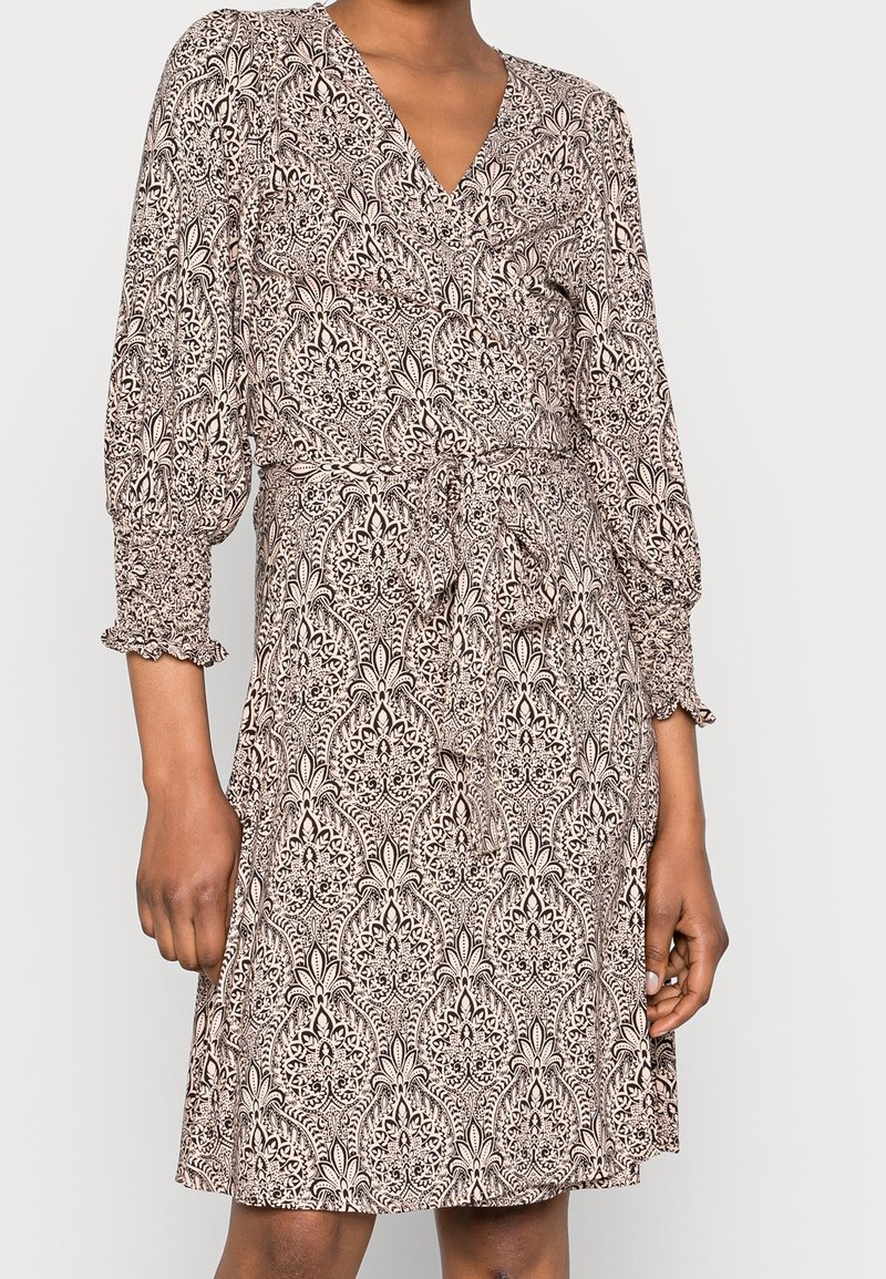 Mujer vistiendo un vestido envolvente beige hasta la rodilla con un intrincado patrón floral negro y mangas tres cuartos recogidas, atado en la cintura.