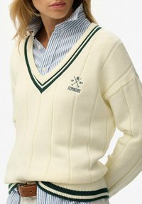 Cremefarbener V-Ausschnitt-Pullover mit grünen Streifen am Kragen, gerippter Textur und einem Logo auf der Brust, kombiniert mit einem blau gestreiften Hemd.