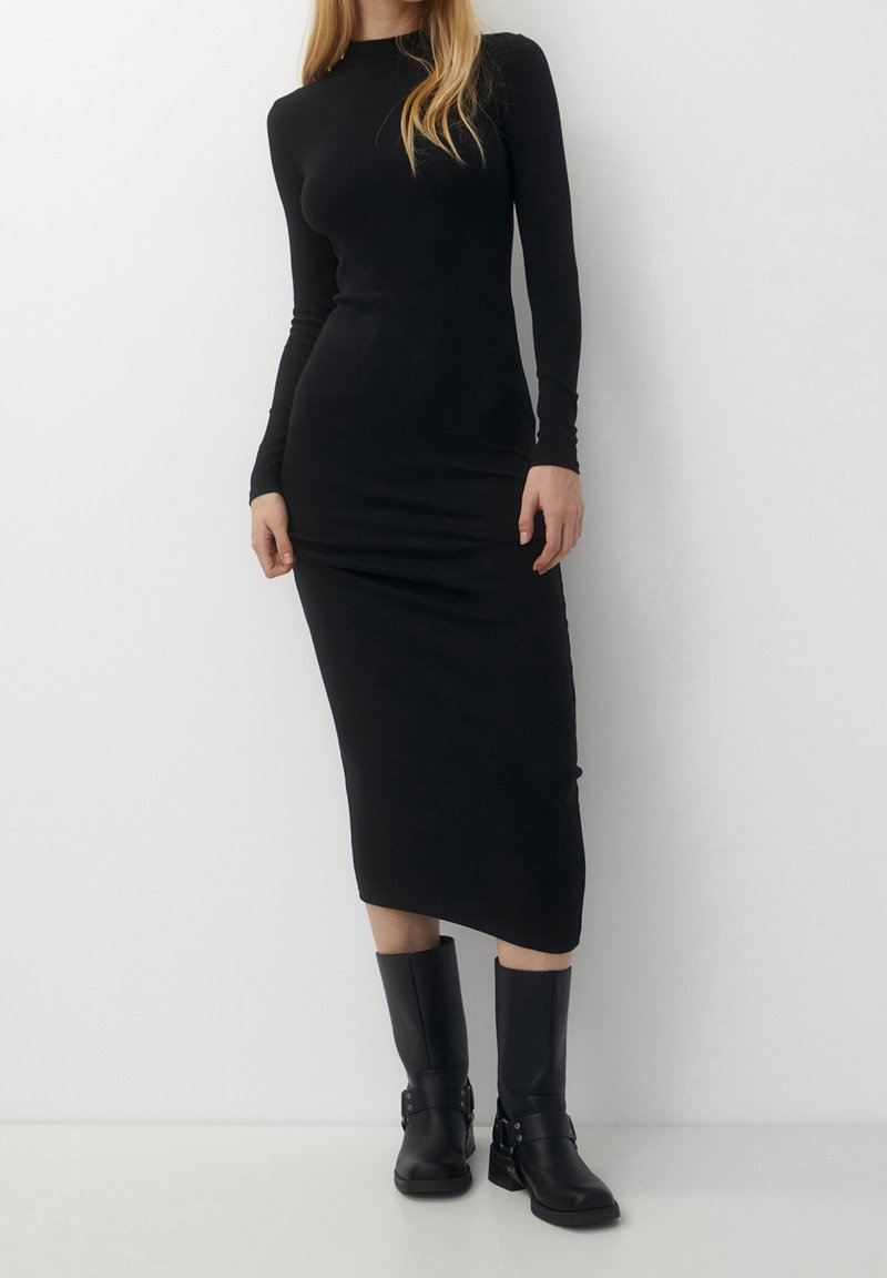 Schwarzes langärmliges Kleid aus dehnbarem Stoff, mit einer figurbetonten Silhouette und knielangem Saum. Getragen mit knielangen schwarzen Stiefeln.