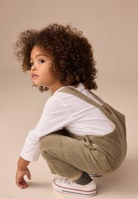 Salopette en velours côtelé olive, avec des bretelles réglables et des accents de boutons, associée à une chemise blanche à manches longues et des baskets blanches.
