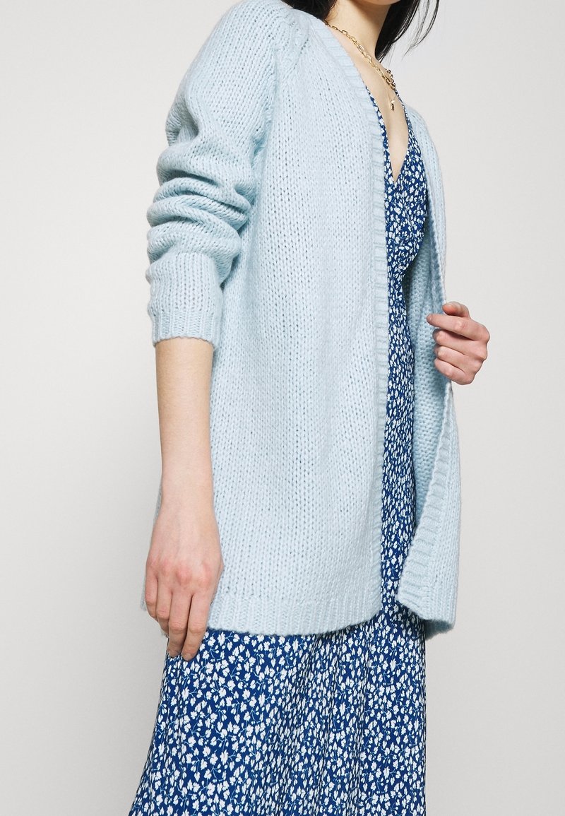 Gros plan d'une personne portant un cardigan tricoté bleu clair sur une robe bleue à fleurs, montrant le milieu du corps et les détails des mains.