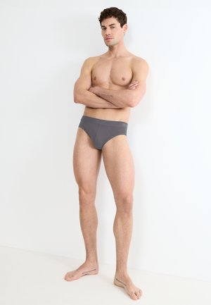 Slip in cotone grigio con vestibilità aderente, dotati di una vita morbida. Il modello è in piedi a piedi nudi, con le braccia incrociate, contro uno sfondo neutro.