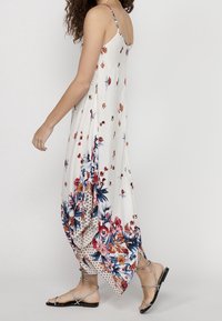 Femme portant une robe longue blanche à fleurs avec des bretelles fines et des sandales plates à lanières, marchant devant un fond uni.