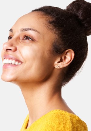 Goldene kreisförmige Steckerohrringe im durchstochenen Ohr mit einer glatten Oberfläche. Die Trägerin hat dunkles, lockiges Haar zu einem Dutt frisiert und trägt ein gelbes Strickoberteil.