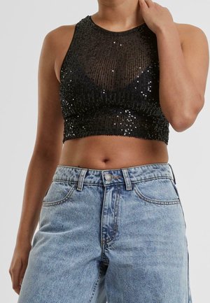 Kobieta w czarnym cekinowym krótkim topie i jasnoniebieskich dżinsach z wysokim stanem, jedną ręką dotykająca szyi.