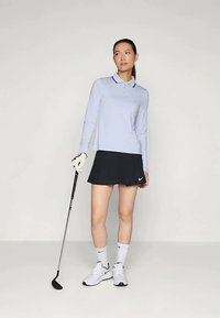 Gai zils garām piedurknēm polo krekls ar apkakli, melna golfa svārki, balti sporta apavi un golfa nūja. Gluda auduma virsma ar kontrastējošām akcentiem.