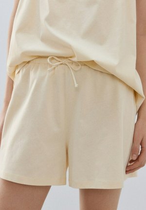 Svetlo bež priložene hlače s elastičnim pasom in zavezanim vezalko, nošene z ujemajočo se sproščeno zgornjo majico, prikazan del ene roke.