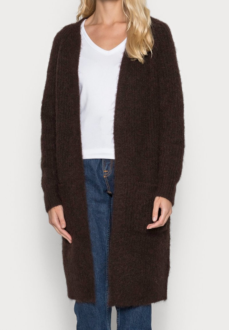 Blonde woman wearing a long, dark brown fuzzy cardigan over a white V-neck shirt and blue jeans, standing against a plain background.