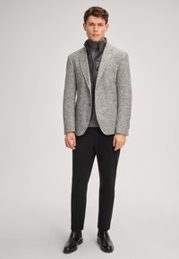 Grau karierter Tweed-Blazer mit Kerbkragen, darüber eine schwarze, gesteppte Weste mit Reißverschluss und ein grauer Rollkragenpullover, kombiniert mit schwarzer Hose und Stiefeln.