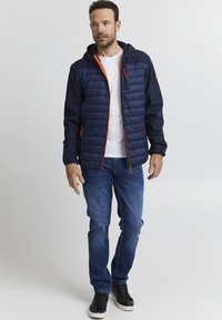 Marineblå polstret jakke med orange detaljer, lommer med glidelås, og hette. Båret over en hvit T-skjorte og sammen med blå jeans og svarte sneakers.