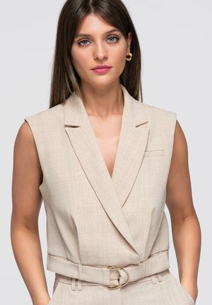 Frau mit glatten braunen Haaren trägt einen ärmellosen beigen Blazer mit Gürtel über passende Hosen vor einem schlichten hellen Hintergrund.