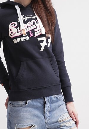 Kobieta w granatowej bluzie z kapturem z białymi sznurkami i nadrukiem, w połączeniu z jasnoniebieskimi, podartymi jeansowymi szortami.